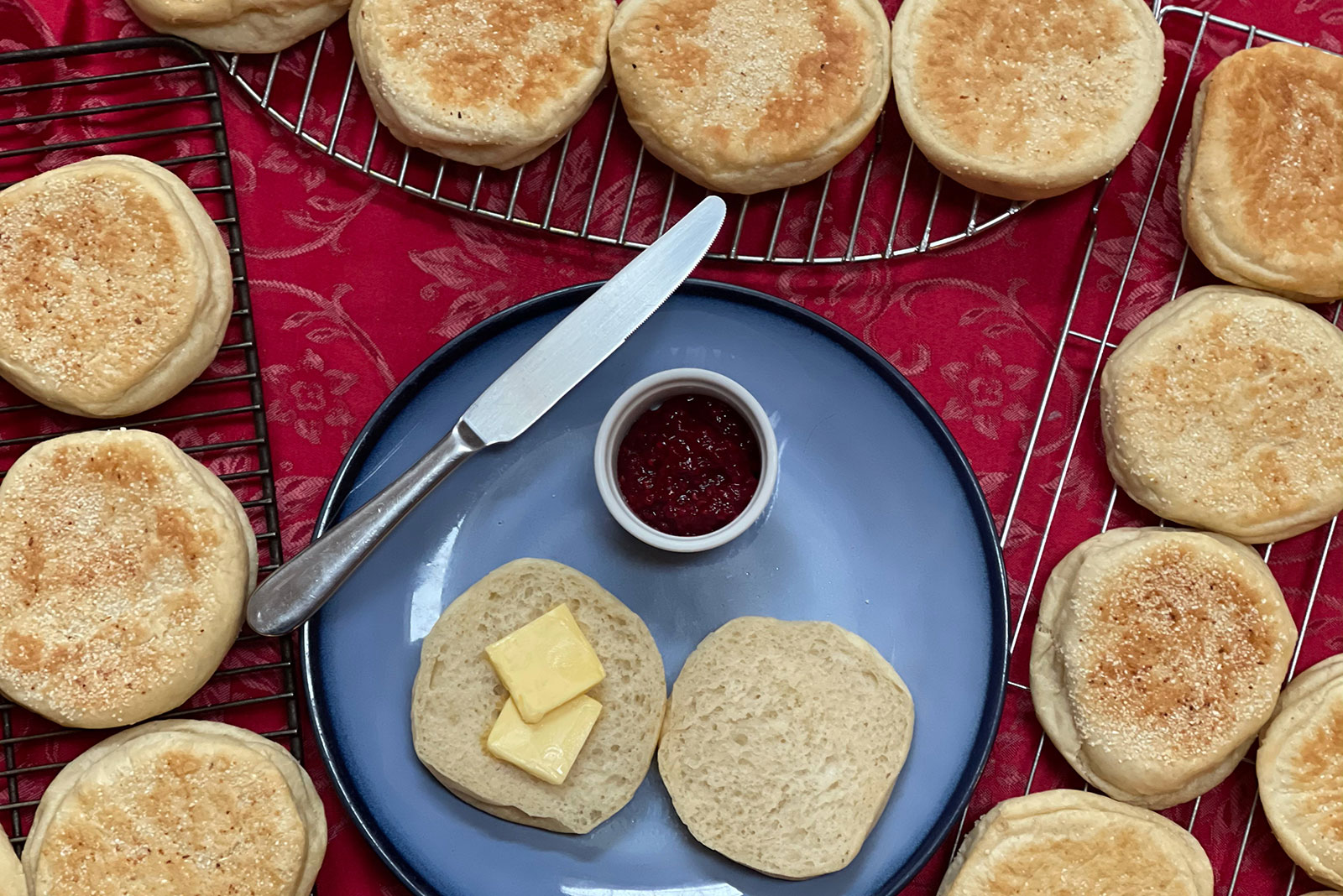 English muffins cooling on racks around a blue plate with jam, butter, and a knife