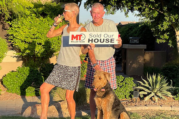 Two people standing with their dog in the sun holding a sold sign
